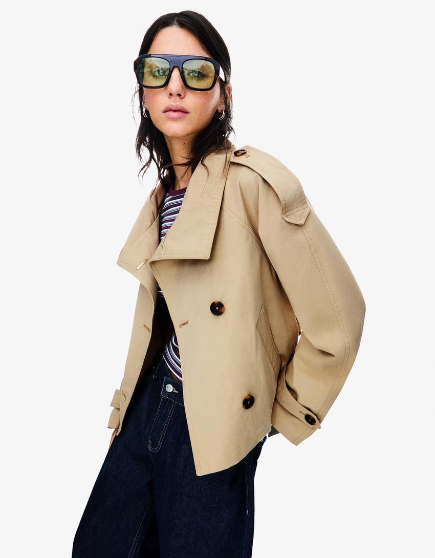 frau trägt beige cropped trenchcoat mit epauletten und doppelknöpfen